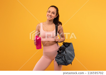 A cheerful young woman dressed in a pink sports bra and leggings stands confidently against a vivid yellow backdrop, holding a water bottle in one hand and slinging a gym bag over her shoulder A cheerful young woman dressed in a pink sports bra and leggings stands confidently against a vivid yellow backdrop, holding a water bottle in one hand and slinging a gym bag over her shoulder 116595702