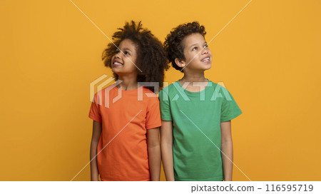 Two joyful African American children are standing side by side, one wearing an orange shirt and the other in green, both gazing upwards with happy expressions on their faces 116595719