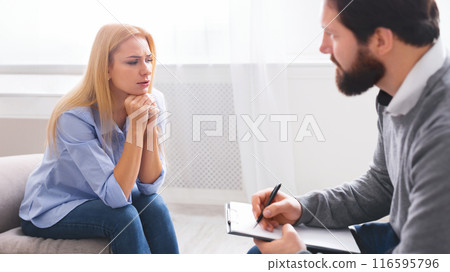 A thoughtful woman is engaged in a serious conversation with a man therapist who is taking notes. They are seated in a well-lit room with a neutral color scheme, indicating a professional setting 116595796