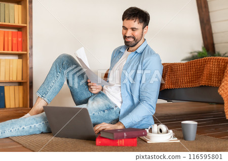 A cheerful man in casual attire sits on the floor with his laptop open and paperwork in hand, sitting area features a coffee cup, headphones, and books, suggesting a relaxing work-from-home setup A cheerful man in casual attire sits on the floor with his laptop open and paperwork in hand, sitting area features a coffee cup, headphones, and books, suggesting a relaxing work-from-home setup 116595801