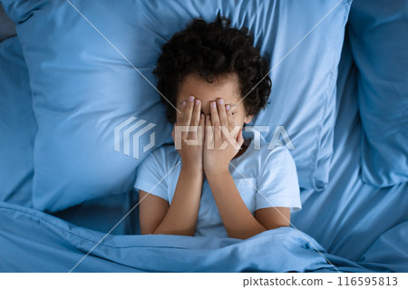 African American young boy child is lying on a bed, with his hands covering his face. He appears to be deep in thought or experiencing emotions like sadness or frustration. 116595813