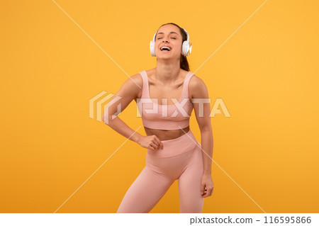 A woman is standing in front of a vibrant yellow background, wearing headphones. She appears focused and engaged, possibly listening to music or a podcast. 116595866