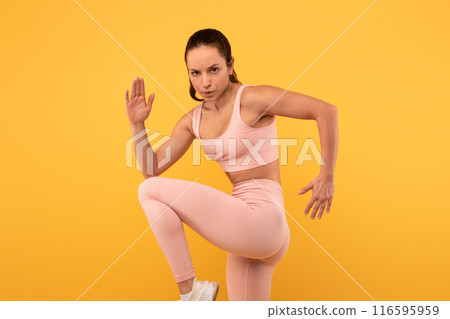 A focused woman is engaging in a physical workout, displaying a motion that suggests a fitness or dance routine, wearing a matching pink sports bra and leggings, vivid yellow backdrop 116595959