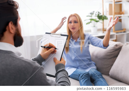 A young woman is seated on a couch, gesturing with her hands as she communicates to man psychologist who is attentively listening and taking notes on a clipboard A young woman is seated on a couch, gesturing with her hands as she communicates to man psychologist who is attentively listening and taking notes on a clipboard 116595983