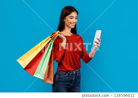 A woman standing holding shopping bags in one hand and a cell phone in the other hand. She appears to be multitasking, possibly checking her phone while shopping, blue background 116596012