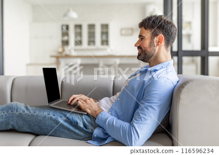 This image shows a man relaxing on a gray sofa, using a laptop. He is wearing jeans and a light blue button-up shirt and is looking at the laptop screen while typing. This image shows a man relaxing on a gray sofa, using a laptop. He is wearing jeans and a light blue button-up shirt and is looking at the laptop screen while typing. 116596234