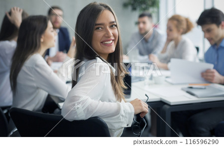 Positive secretary smiling to camera during meeting with colleagues working on background Positive secretary smiling to camera during meeting with colleagues working on background 116596290