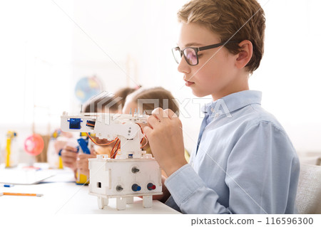 A young boy with glasses is sitting at a table in a classroom setting and is working on building a small, white robotic arm. He is holding a small piece of the robot in his hand. A young boy with glasses is sitting at a table in a classroom setting and is working on building a small, white robotic arm. He is holding a small piece of the robot in his hand. 116596300
