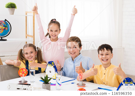 Four young students, two girls and two boys, sit at a table in a classroom setting with thumbs up after successfully completing a STEM activity. Four young students, two girls and two boys, sit at a table in a classroom setting with thumbs up after successfully completing a STEM activity. 116596350
