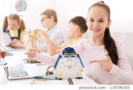 A young girl smiles proudly as she points to a robot she has built in a classroom setting. Other students are visible in the background, suggesting that she is part of a class or group project A young girl smiles proudly as she points to a robot she has built in a classroom setting. Other students are visible in the background, suggesting that she is part of a class or group project 116596365