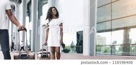 African American couple walks together outside a modern building with large windows, each pulling a suitcase. The couple is dressed casually in summer clothes, copy space African American couple walks together outside a modern building with large windows, each pulling a suitcase. The couple is dressed casually in summer clothes, copy space 116596406