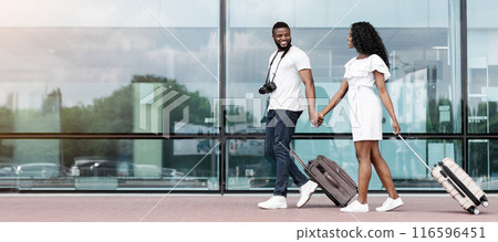 Black couple walks together through the airport terminal, holding hands and pulling their luggage behind them. They both appear to be smiling and happy, copy space 116596451