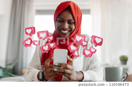 African American woman wearing a red hijab smiles while looking at her smartphone, surrounded by pink heart icons representing social media likes. She is sitting at a table in a bright, modern room. 116596473