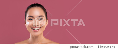 Cheerful attractive topless young asian woman with moisturizer cream on her pretty face, testing new beauty product, smiling at camera, purple studio background, copy space. Face care concept 116596474