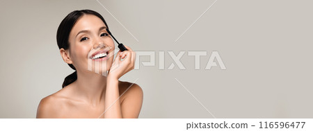 Pretty half-naked young woman applying makeup, holding mascara and smiling at camera, isolated on grey studio background, copy space 116596477