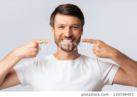 A man with a beard smiles widely and points to his teeth with both index fingers. He is wearing a white t-shirt and stands against a white background. 116596481