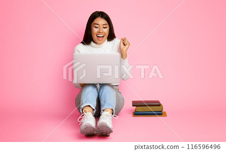 A young Asian woman sits on a pink background, radiating joy as she uses a laptop. Her hands are raised in celebration, expressing triumph. A stack of books sits beside her, studious setting. 116596496