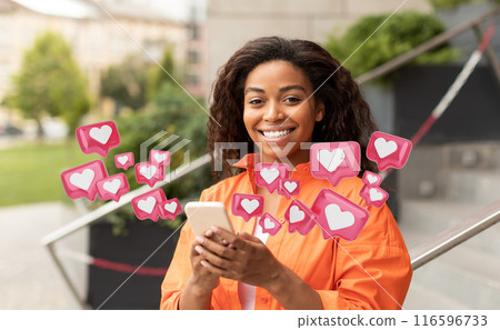 African American young woman in an orange shirt smiles at the camera while holding a smartphone and looking at notifications. Surrounding her are digital heart icons representing social media likes. 116596733