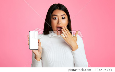 Asian young woman with long dark hair, wearing a white sweater, standing in front of a pink background. She is holding a smartphone with a blank screen in her left hand and covering her mouth 116596755