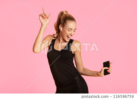 Workout playlist. Young woman holding smartphone dancing listening to music on yellow studio background. Empty space 116596759