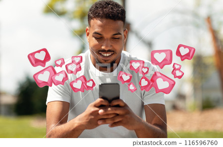 A young black man is sitting in a park, smiling while looking at his smartphone. He is holding the phone with both hands, and there are multiple heart-shaped icons floating around him 116596760