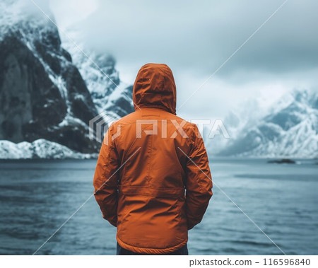 Man orange jacket back view stands rocky shore gazing cold misty fjord snow mountains Nordic winter 116596840