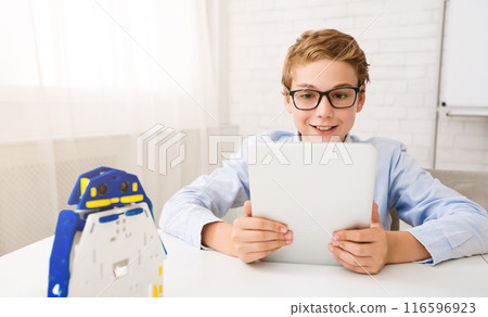A young boy with glasses sits at a desk and uses a tablet. He looks intently at the screen, smiling. There is a small robot toy on the desk in front of him. A young boy with glasses sits at a desk and uses a tablet. He looks intently at the screen, smiling. There is a small robot toy on the desk in front of him. 116596923