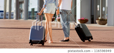 Traveling Together Concept. Rear view of two tourists walking in or out airport, carrying suitcases, holding hands Traveling Together Concept. Rear view of two tourists walking in or out airport, carrying suitcases, holding hands 116596949