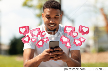 A young black man wearing a white t-shirt uses his smartphone, as digital pink hearts with white centers surround him, suggesting he is receiving many likes or positive feedback on his phone. A young black man wearing a white t-shirt uses his smartphone, as digital pink hearts with white centers surround him, suggesting he is receiving many likes or positive feedback on his phone. 116597039