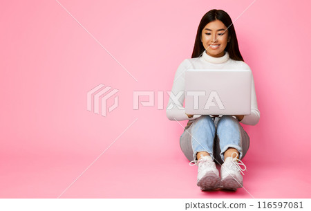 Asian young woman with long dark hair is sitting on a pink background, looking down at a laptop. Woman is smiling and appears to be working or browsing the internet, copy space 116597081