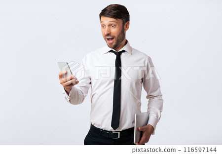 Good News. Excited Man Holding Smartphone Standing With Opened Mouth In Surprise On White Background In Studio. Empty Space 116597144