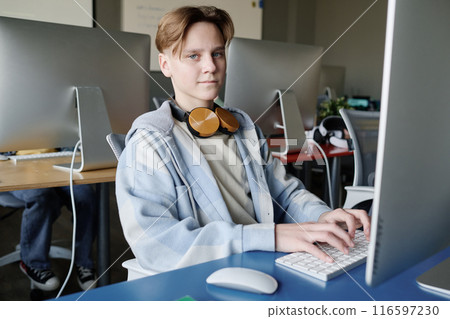 Medium shot of modern teen boy wearing casual clothes with headphones sitting at desk in classroom typing on computer Medium shot of modern teen boy wearing casual clothes with headphones sitting at desk in classroom typing on computer 116597230