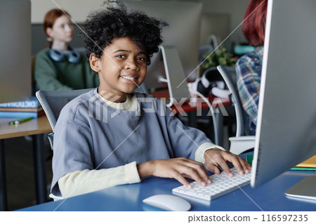 Medium shot of cheerful African American boy sitting at desk in modern classroom smiling at camera while typing on computer 116597235
