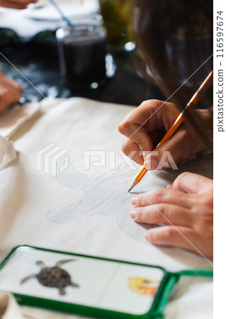 Girl copying the design of a turtle from the phone on a piece of clothing with a pencil. Vertical Girl copying the design of a turtle from the phone on a piece of clothing with a pencil. Vertical 116597674