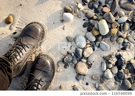 Legs of a standing female in rocker boots on beach sand with various rocks and pebbles in sunset light 116598099