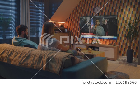 Couple watches news about United States elections on TV screen sitting on sofa Couple watches news about United States elections on TV screen sitting on sofa 116598566