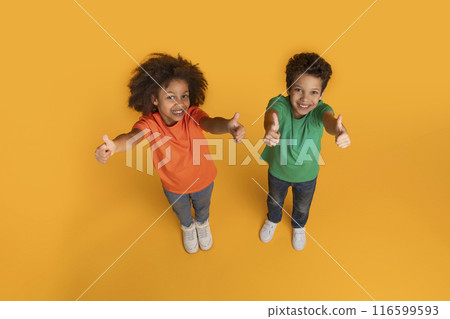 Two African American children, a boy and a girl, are enthusiastically giving thumbs up gestures against a vibrant orange background. Their happy expressions convey approval and positivity, top view 116599593