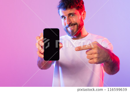 A cheerful man with a beard, wearing a white t-shirt, is excitedly pointing at a smartphone blank screen while standing against a vibrant pink and purple neon background. 116599639