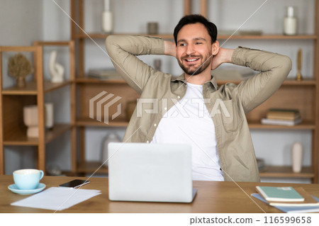 A young man with a cheerful expression sits back at his desk in a well-lit, contemporary office setting, hands clasped behind his head. In front of him is an open laptop, alongside a cup of coffee 116599658