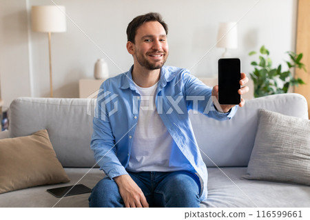 A man is seated on a couch, holding a smartphone in his hand. He appears focused on the screen, possibly browsing or texting. The room is cozy, with a soft light illuminating the scene. A man is seated on a couch, holding a smartphone in his hand. He appears focused on the screen, possibly browsing or texting. The room is cozy, with a soft light illuminating the scene. 116599661