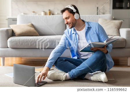 A cheerful man sits comfortably on the floor against a couch, wearing headphones and a blue shirt, engrossed in a book while simultaneously working or studying on a laptop A cheerful man sits comfortably on the floor against a couch, wearing headphones and a blue shirt, engrossed in a book while simultaneously working or studying on a laptop 116599662