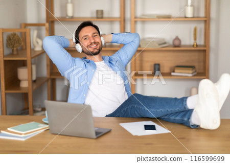 A man is seated at a desk, wearing headphones and working on a laptop placed on his lap. He appears focused and engaged in his work, with various papers and stationery scattered around the desk. A man is seated at a desk, wearing headphones and working on a laptop placed on his lap. He appears focused and engaged in his work, with various papers and stationery scattered around the desk. 116599699