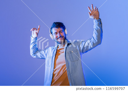 A young man is happily dancing to music while wearing headphones. He appears joyful and relaxed against a vibrant blue background, and it is nighttime. 116599707