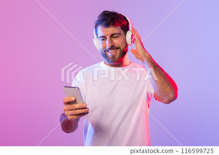 A man is sitting and listening to music through headphones while holding a cell phone in his hand. He appears focused on the music playing and the content on his phone screen. A man is sitting and listening to music through headphones while holding a cell phone in his hand. He appears focused on the music playing and the content on his phone screen. 116599725