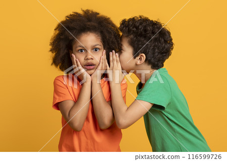 A young African American boy in a green shirt is whispering into the ear of his surprised sister, who is wearing an orange shirt. Her hands are placed on her cheeks, and her expression is astonishment 116599726