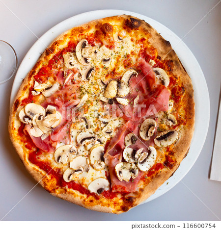 On plate is small round pizza on thin yeast dough with slices of ham and pieces of mushrooms. 116600754