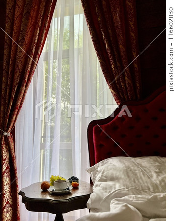 Cup of coffee with fresh fruits on the bedside table in a hotel room with red curtains  116602030