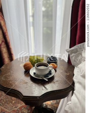 Cup of coffee with fresh fruits on the bedside table in a hotel room with red curtains  116602032