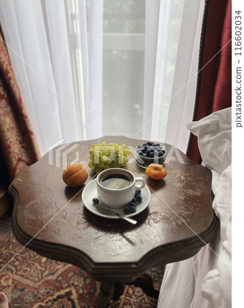 Cup of coffee with fresh fruits on the bedside table in a hotel room with red curtains  116602034
