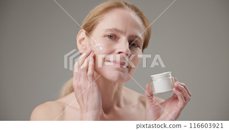 Real people - age, beauty, health and dry skin care concept - beautiful mature Caucasian middle aged woman in her 50s applying moisturizer to her face 116603921